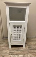 Front view of the white single door bathroom cabinet with woven rattan door panel and the open shelf above.
