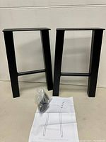 Two black steel trapezoid shaped metal table legs shown side by side on a surface with mounting hardware and installation instructions in front.