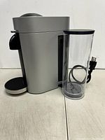 Side view of silver Nespresso VertuoPlus Deluxe machine with transparent water reservoir attached and power cord coiled inside the reservoir.