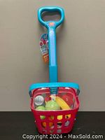 Red and blue rolling shopping basket with handle containing various plastic play foods inside.