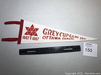 Full view of the 1967 Grey Cup pennant showing red text and design on white felt with ruler for size reference.