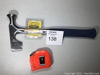 Shows the full hammer with blue handle and metal head that has a hammer face and a claw end; original tag attached at base of hammer head; tape measure also shown but not included.