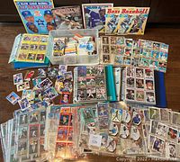 Wide view showing loose cards, binders with baseball card pages, plastic bin with cards, Panini sticker albums, and baseball card magazines.