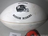 Full side view of the football showing Ottawa Rough Riders helmet logo and multiple faded signatures on white panel.
