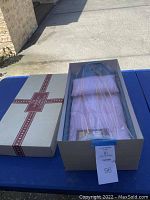 Boxed American Girl Doll bed set on blue table with box lid removed, showing comforter and pillow inside plastic wrapping.