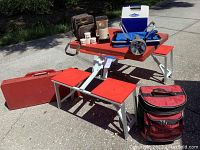 Two portable picnic tables with red tops and attached seats shown folded out on pavement. Several items are placed on top including a small cooler, coffee pot kit, and a large flashlight. Carry cases and a soft cooler are visible nearby.