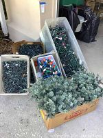 Multiple boxes containing assorted Christmas lights and artificial pine garlands seen outside on concrete floor near a wall.
