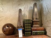Set of two medium brown leather covered obelisks about 18 inches tall, stacked faux book boxes in green and brown colors, and a vintage wooden bowling ball with three visible finger holes on a wood shelf against a stone wall.