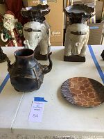 Lot showing two white elephant plant stands with dark saddle tops, dark clay elephant pot, and painted giraffe plate on table.