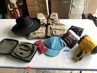 Lot items arranged on a white table. Included are black felt western hat, beige life vest, padded Oakley sunglasses case, blue women's cap with pink bow, decorative hair combs, and yellow Garrit spotlight.