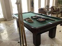 Full view of Olhausen pool table with green felt, wooden frame, pool cues leaning and accessories on top