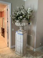 Full view of plant stand with urn and floral arrangement in a corner near doorway and wall vent.