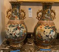 Pair of Satsuma style vases with detailed hand painted crane and floral designs, gilded dragon handles, and scalloped patterned rims; placed on marble surface.