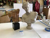 Photo showing two busts on a table: a male torso with no head or arms on a black base, and a female bust with head and partial shoulders.
