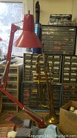 Trumpet hanging on a wall beside a red adjustable lamp, near a multi-drawer plastic organizer containing small parts and tools.