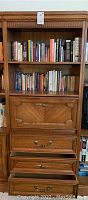 Full view of wooden bookcase showing two open shelves filled with books and three lower drawers with decorative paneling and metallic handles, top drawer being a drop front panel.