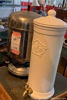 Photo shows stainless steel coffee urn and white ceramic water filter together on a wooden surface, highlighting overall size and condition.