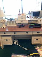 Full view of wooden desk with brown surface, off-white drawers, and cubby with multiple compartments, white ornate lamp with shade, small TV, and some miscellaneous items on top.