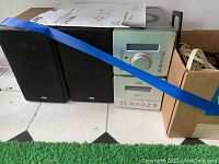 Photo showing JVC main component system unit, separate CD player, and two black JVC speakers side by side on floor with a paper manual/instructions on top of one speaker.