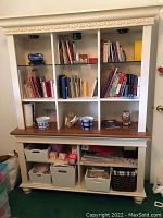 Front view of the white wood bookshelf filled with books, two blue and white ceramic pots, woven basket, bonded notebooks, and wooden bins with stationery items.