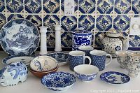 Full assortment of blue and white china including teapot, bowls, mugs, elephant bank, rooster bowls, and candlesticks arranged on a kitchen counter with decorative blue and white tiles as background.