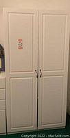 Front view of closed tall white cabinet showing two paneled doors and metal handles with a price sticker.