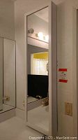 Front view of tall white jewelry organizer cabinet with full-length mirror door hanging on wall above bathroom counter with sink and light switch to right.