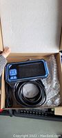 Open cardboard box with digital inspection endoscope device visible inside with attached cable and screen with control buttons.