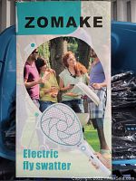 Front of the box showing the brand name Zomake and product name Electric Fly Swatter with a photo of people socializing outdoors and a large image of the electric swatter device.