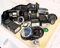 Photo shows a wide assortment of camera flashes, lens adapters, lens hoods, and light meter on a white background.