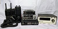 Wide view of full lot showing two black vintage walkie talkies, multiple black and gray CB radios, coiled microphone, and cream-colored Morse Code unit.