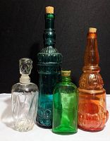 Four decanter bottles in clear, blue-green, green, and amber glass arranged on white surface against black background, showing their shapes and stoppers.