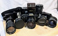 Lot overview showing three camera flashes, Canon camera with lens, and multiple lenses with cases on wooden surface.