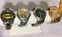 Four men's watches arranged on white display stand including Batman logo, Fossil, black face watch and rose gold tone watch with leather band.