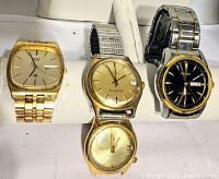 Four men's watches displayed on a white padded holder showing a gold-tone Timex, gold-tone Seiko, black dial Pulsar, and another gold-tone Accutron watch without the band.