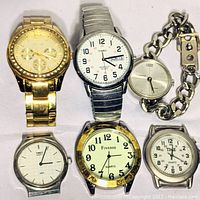 Top-down view of six men's metal watches arranged in two rows on white surface, showing faces and bands (some missing bands)