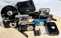 Seven Nikon Coolpix cameras in various colors and models displayed with Nikon Software Suite CD, camera case, batteries and charger on wooden surface.