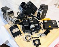Photo showing multiple vintage cameras and accessories on a wooden surface including Bell & Howell Super 8 model, Minolta 110 Zoom SLR, lens covers, and an extended viewer
