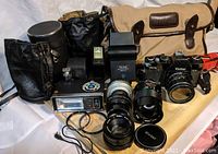 Overall lot viewed from front showing cameras, lenses, flash, cases and carrying bag arranged on wooden surface with cloth backdrop.