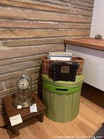 Overall photo of the Kundo anniversary clock on a small wooden stool beside the green round storage container and wicker basket holding picture frames and small chest box.