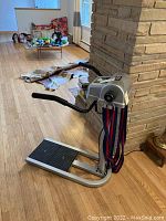 Full view of the Beauty Belt Massager showing the foot platform, side handles, main unit and belts hanging from it, positioned next to a stone wall.