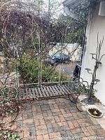 Outdoor metal bench on brick patio surrounded by greenery, showing side armrest with scroll detail