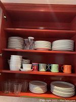 Full view of kitchen cabinet shelves showing stacked white plates, bowls, colorful mugs, clear glasses, and clear canisters