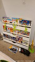 Photo shows a white wooden book rack filled with various children's books and learning workbooks, with three metal green monkey hangers attached to the rack. A black Lego Creator car toy and yellow Money Gobbler giraffe toy are on the floor next to the rack.