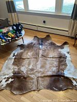 Full view of natural cowhide rug laid flat on wooden floor under natural light, showing brown and white spots.