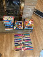 Photo shows multiple board games and puzzles stacked in three piles on a fireplace hearth, with children's books laid face up on the floor in front of the hearth.
