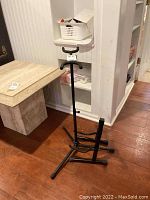 Two black metal adjustable instrument stands on hardwood floor beside shelves and furniture, showing structure and height adjustment levers.