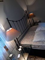 Pair of metal table lamps shown on bedside tables next to bed frame. Each lamp has beige fabric shade and twisted metal base.