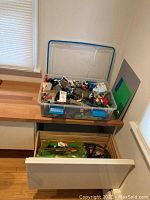 Photo showing clear plastic container with assorted Lego bricks inside on wooden counter and Beyblade arena in drawer beneath.