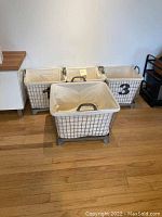 Four metal canvas baskets with beige fabric liners arranged on wooden floor